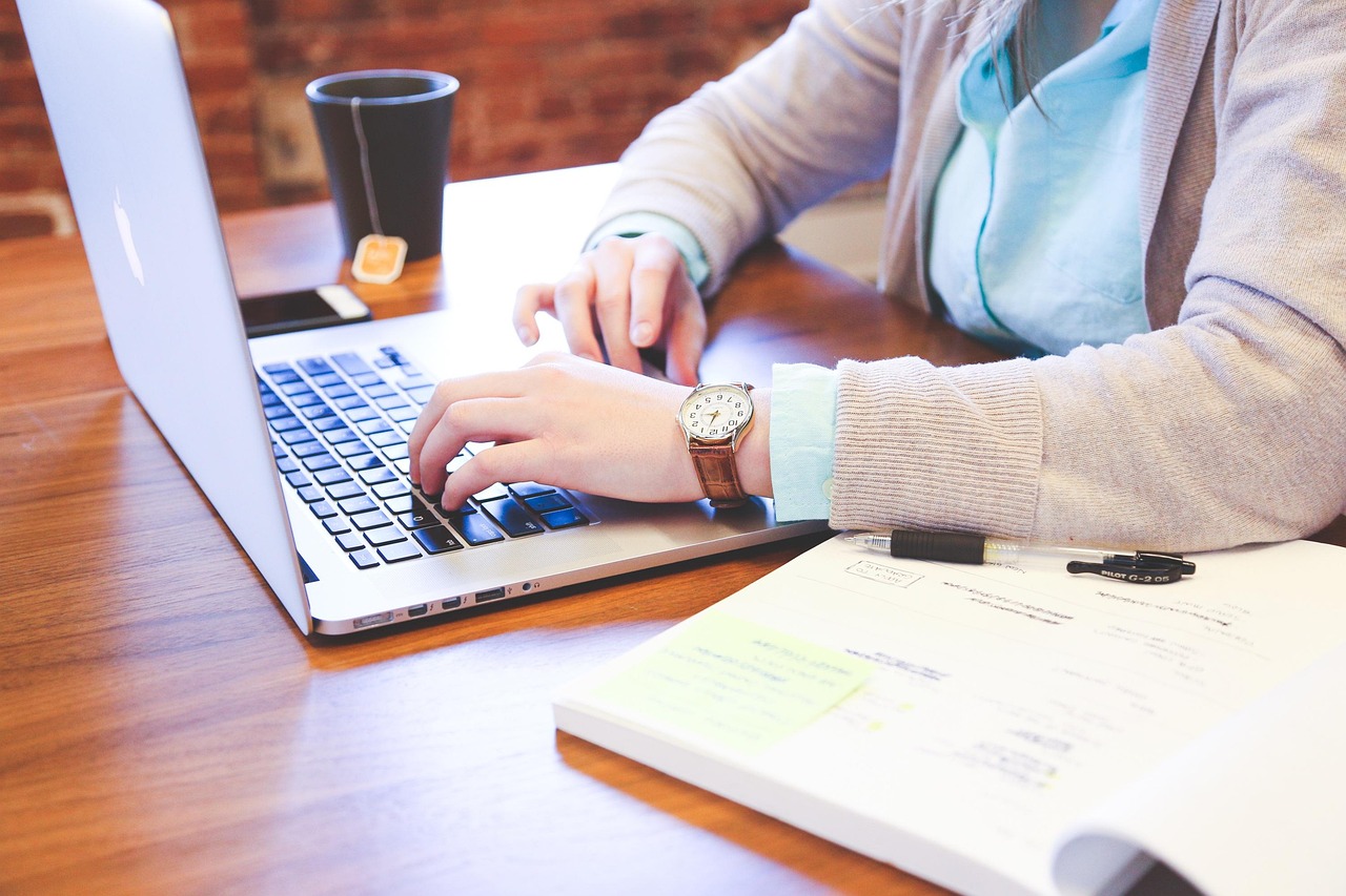 Osoba pracująca na laptopie przy biurku z notatnikiem i kawą